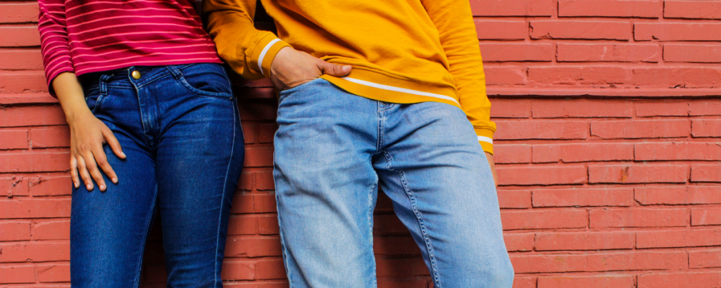 Two people standing against a red brick background with jeans and colorful shirts